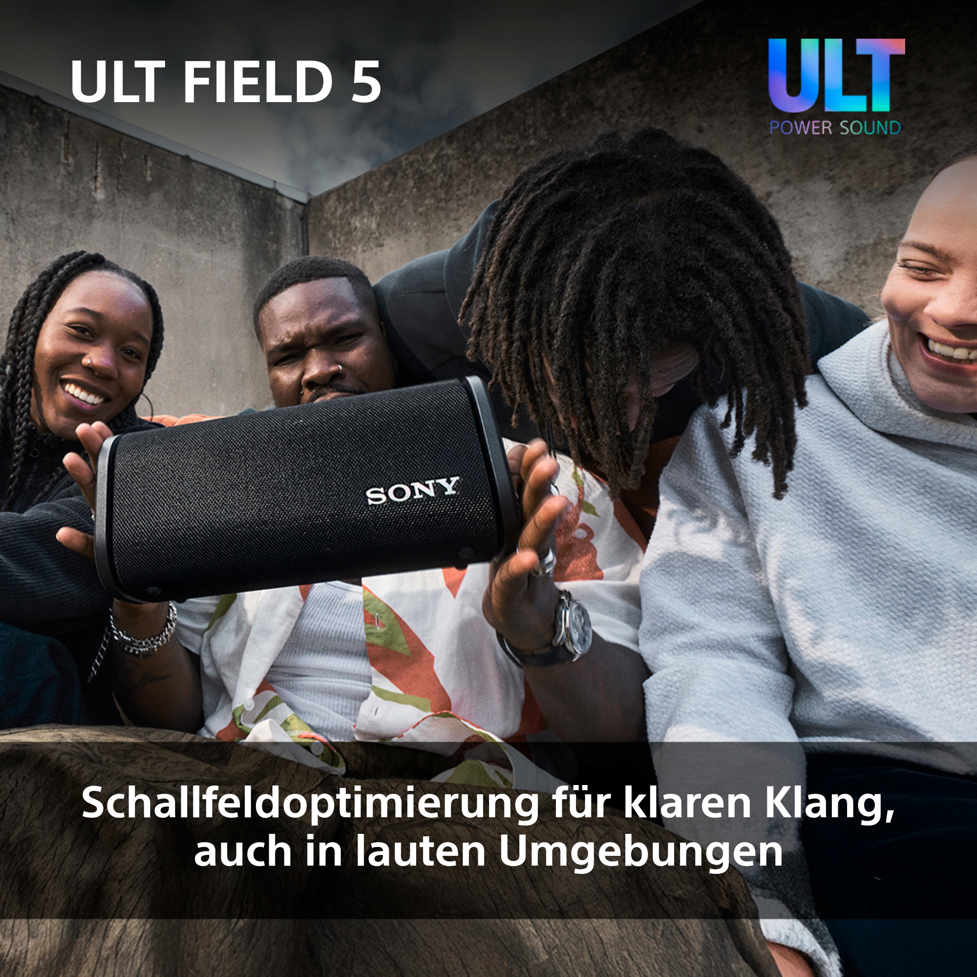 Gruppe von Leuten um einen schwarzen Lautsprecher, lächelnd und interagierend, mit grauem Hintergrund.