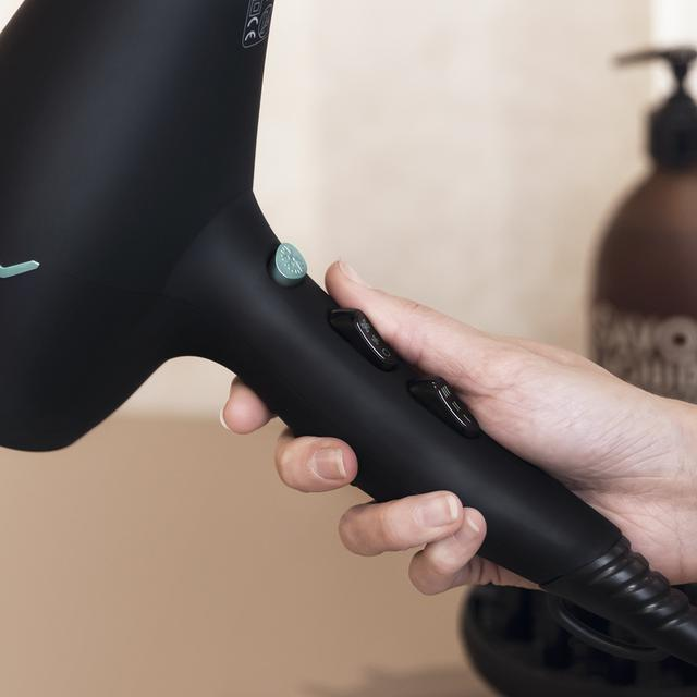 Dłoń trzymająca czarną suszarkę do włosów, obok blatu łazienkowego i butelki.