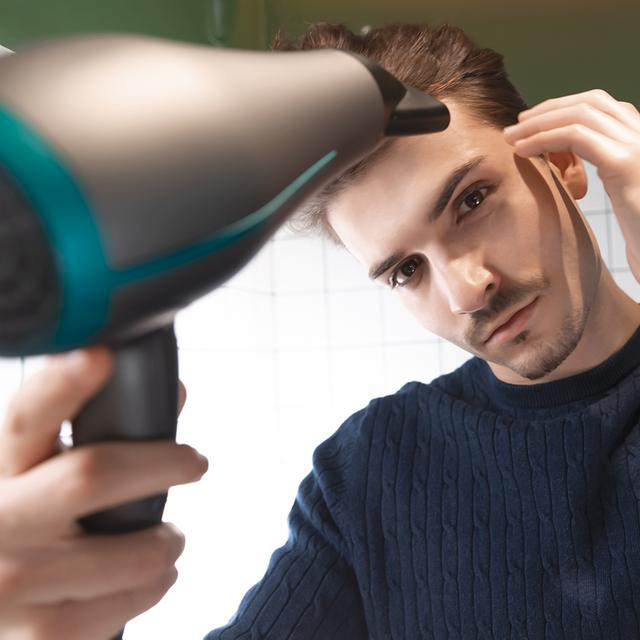 Mężczyzna suszy włosy suszarką. Patrzy w kamerę.