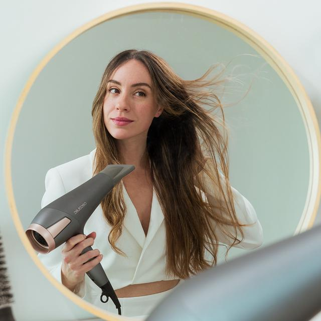 Kobieta spogląda w lustro, trzymając szarą i różowo-złotą suszarkę do włosów.