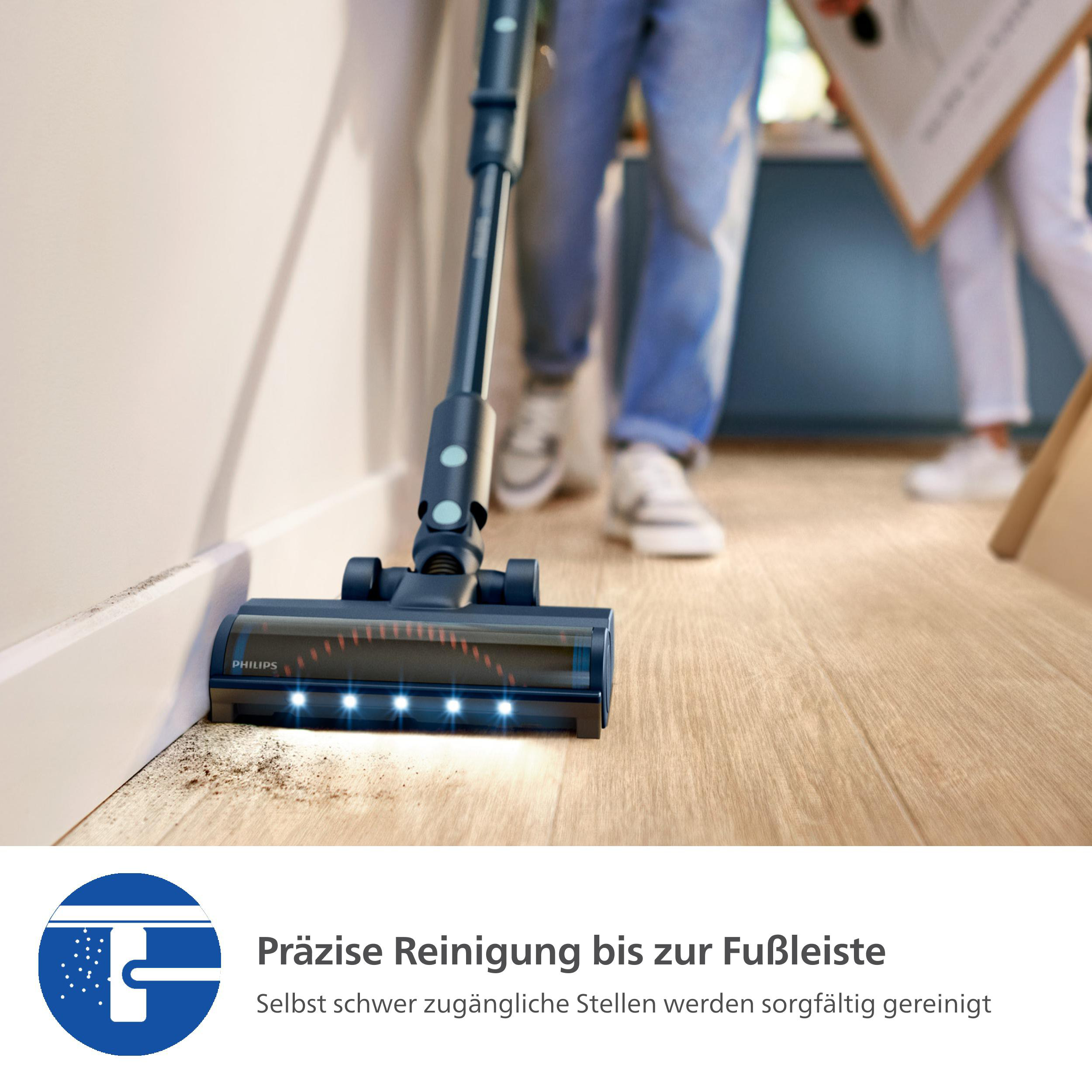Staubsauger reinigt Holzboden nahe einer Wand. Eine Person geht im Hintergrund.