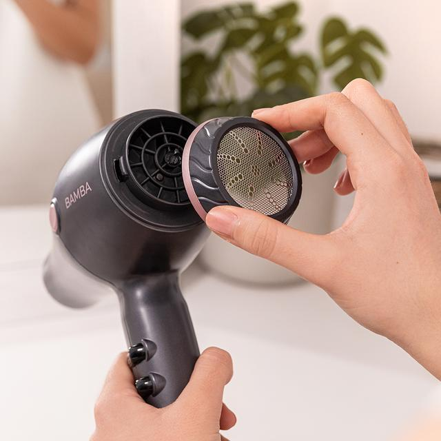 Osoba wyjmująca filtr z czarno-szarej suszarki do włosów. Różowe akcenty. Białe tło.