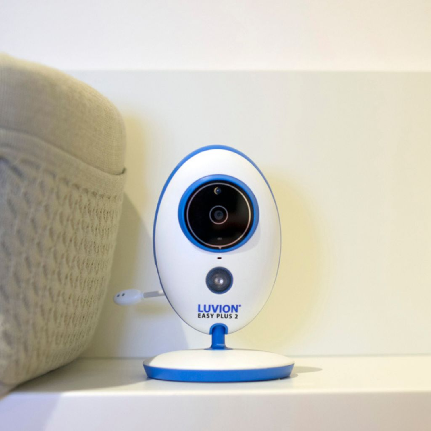 Een witte en blauwe babyfoon staat op een plank. De monitor heeft een cameralens en een knop.
