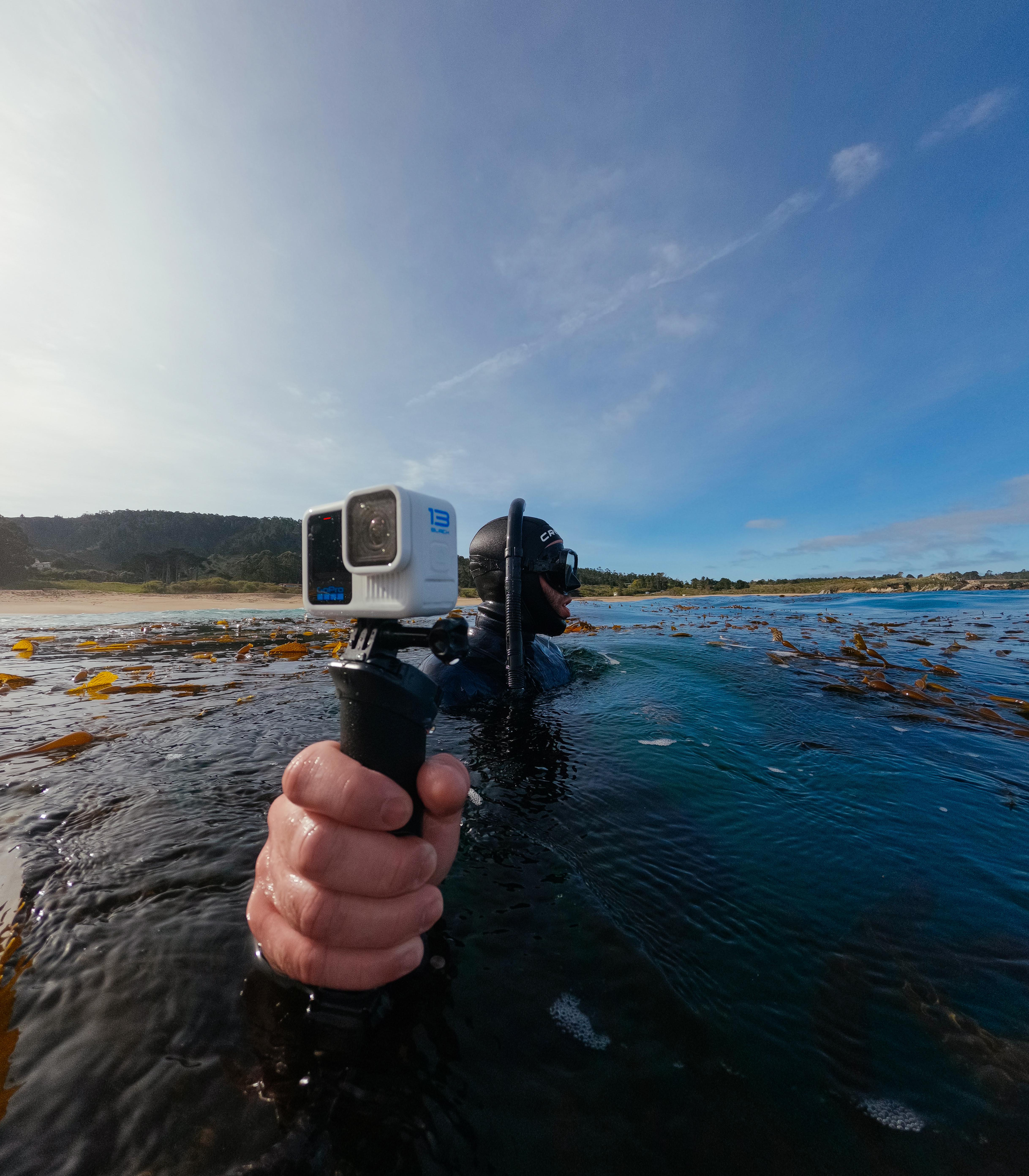 Person im Taucheranzug hält eine Kamera im Wasser, mit blauem Himmel.