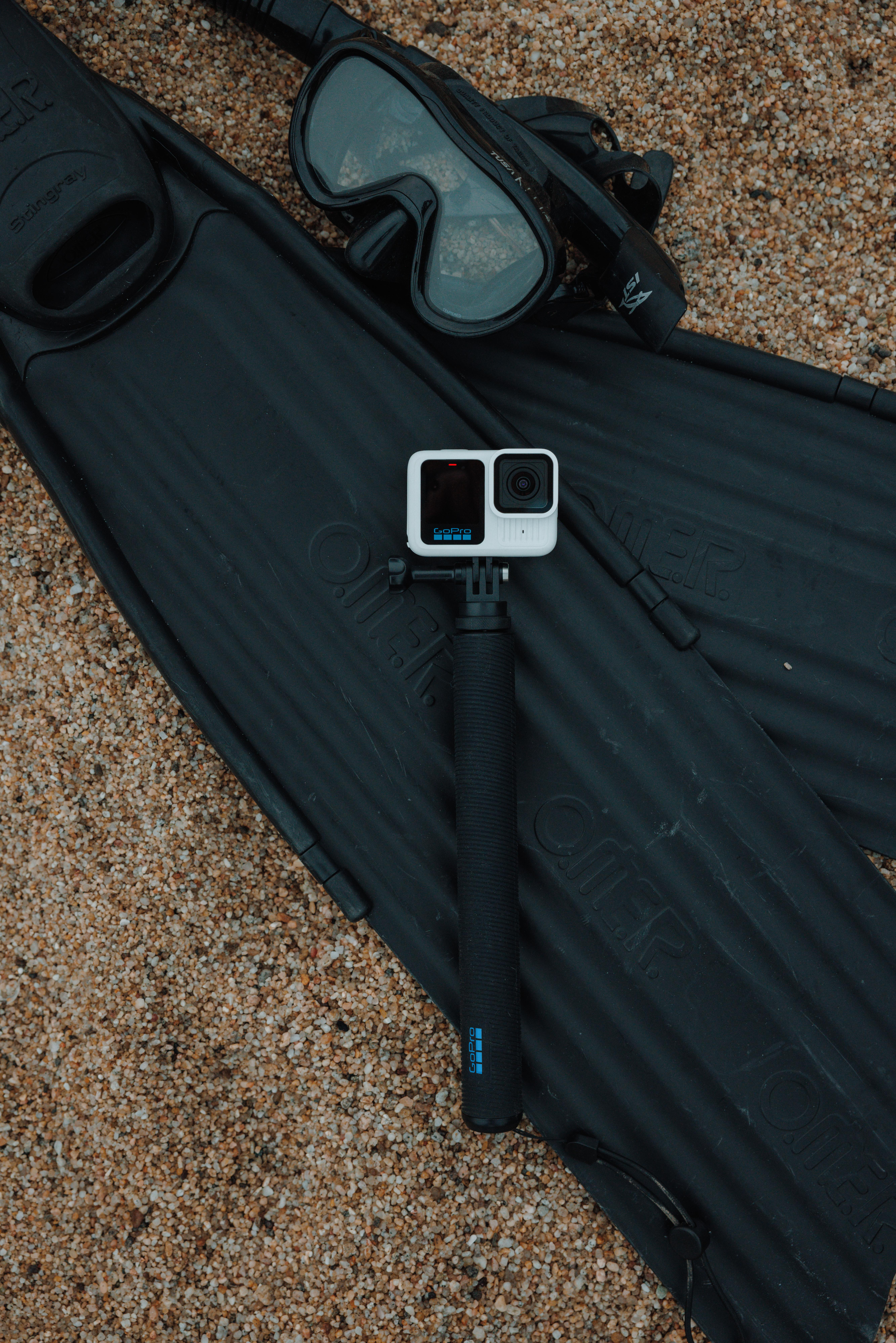 GoPro-Kamera auf einem Stab, Flossen und eine Tauchmaske auf Sand.