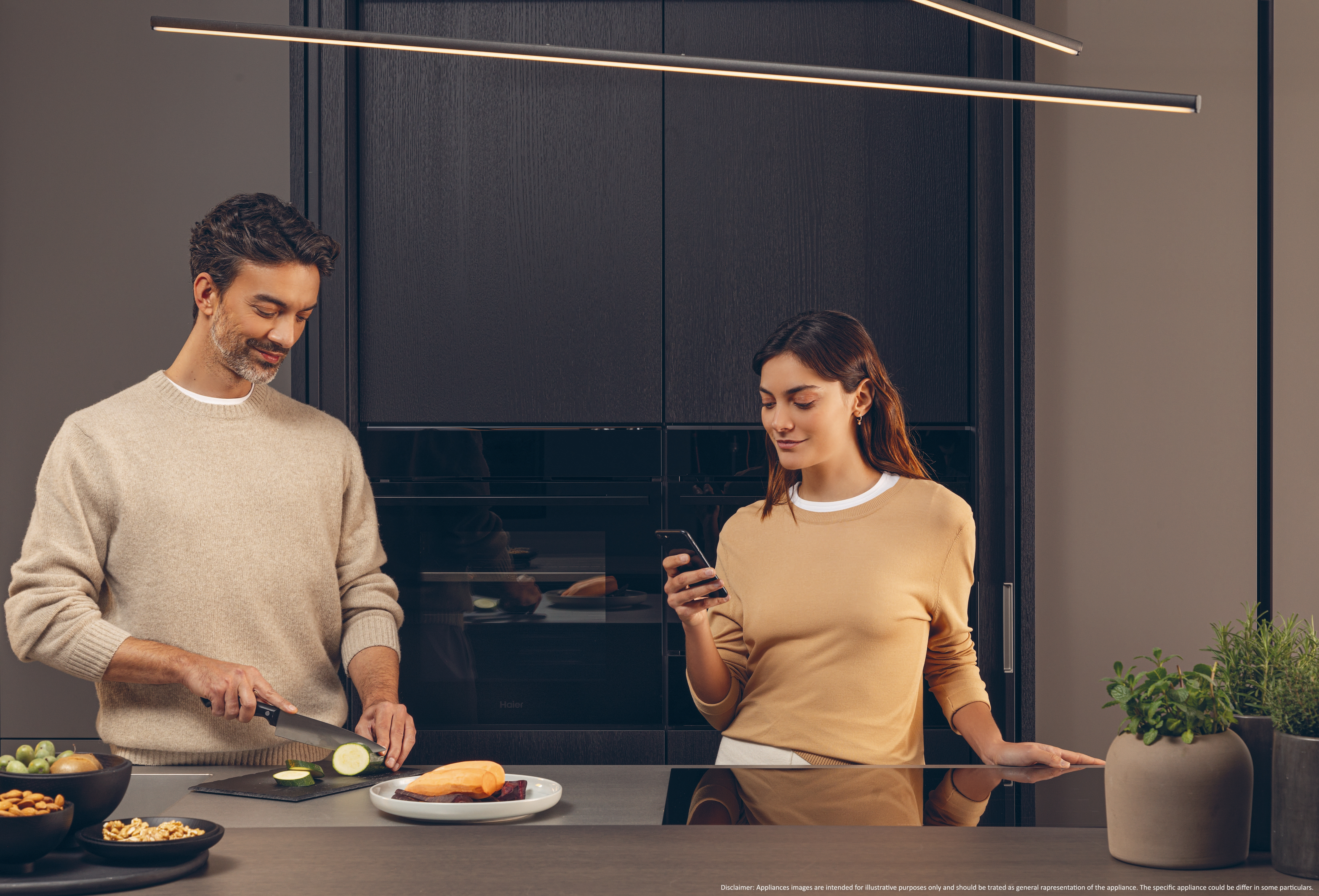 Una pareja en una cocina moderna; uno corta verduras y el otro usa un teléfono.