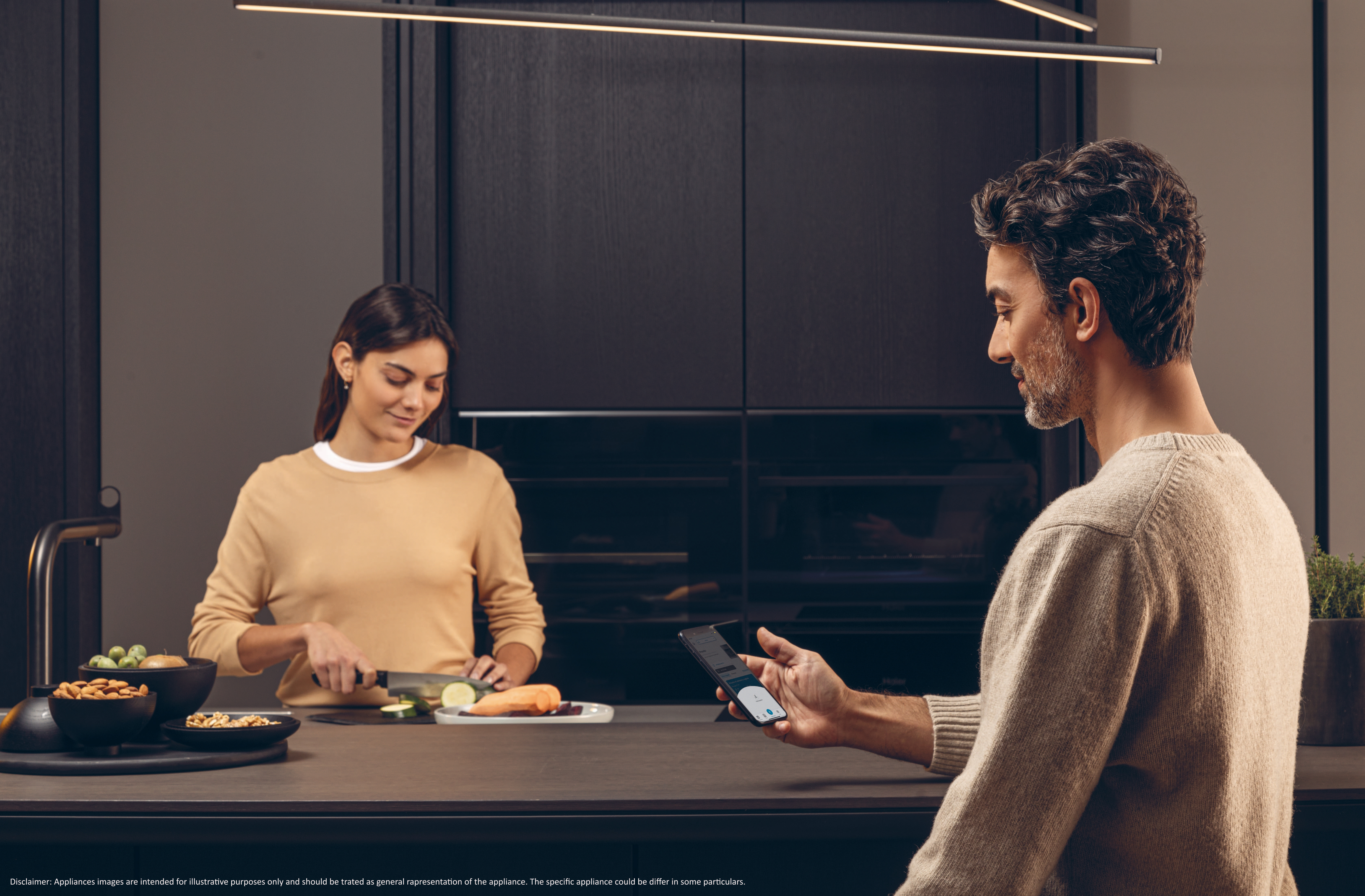 Una mujer corta verduras en una cocina. Un hombre mira un teléfono. Armarios oscuros al fondo.