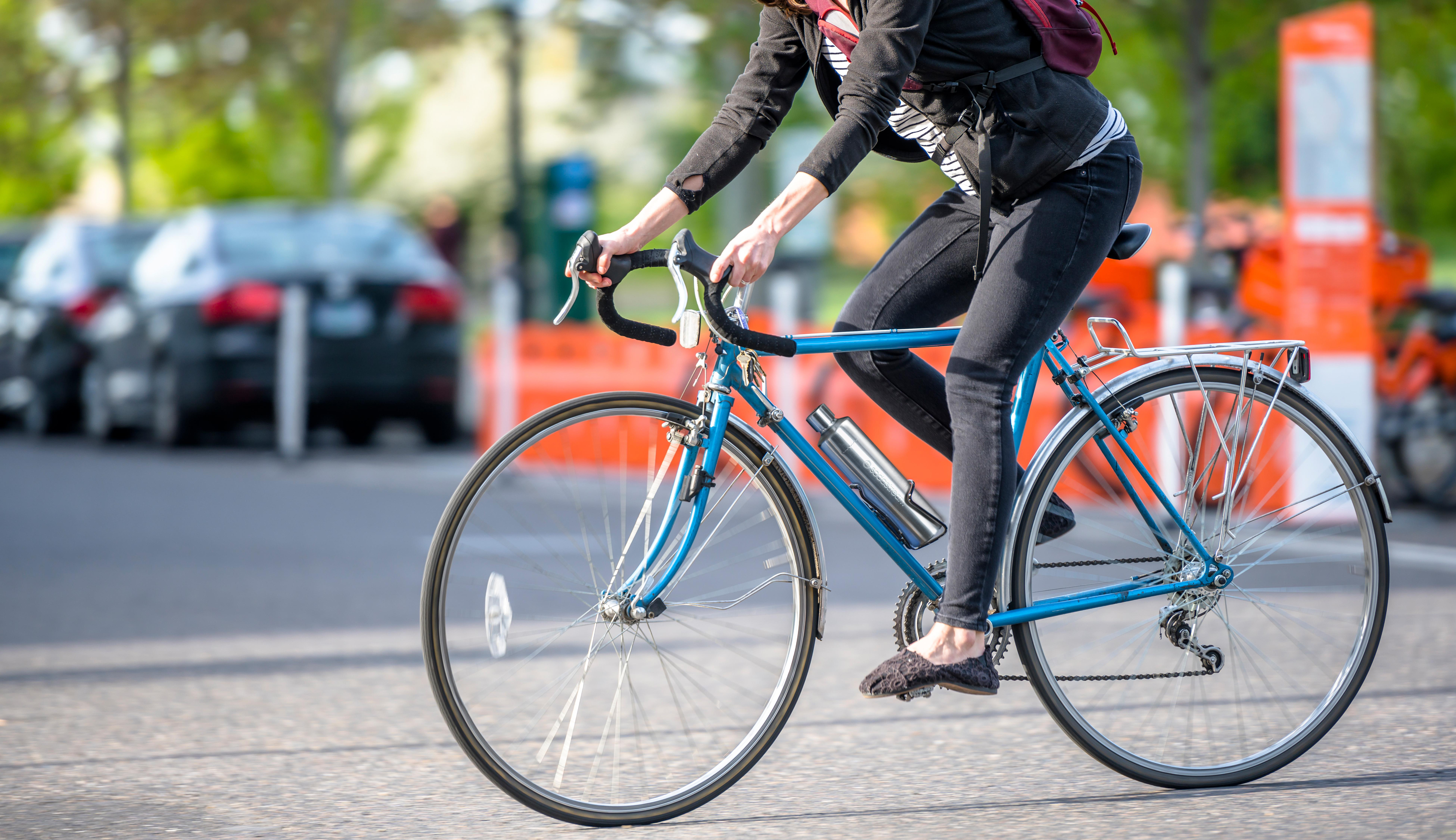 Person fährt blaues Fahrrad auf Straße. Autos und orangene Kegel im Hintergrund.