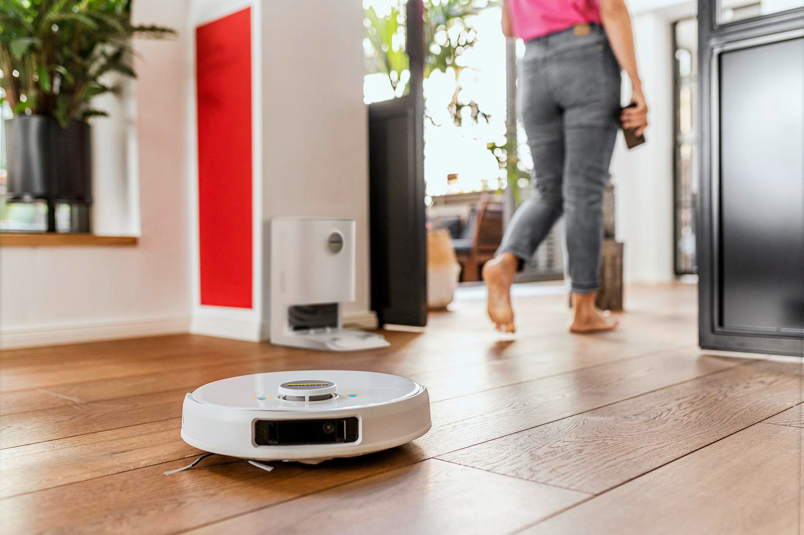 Roboter-Staubsauger auf Holzboden, Frau im Hintergrund.