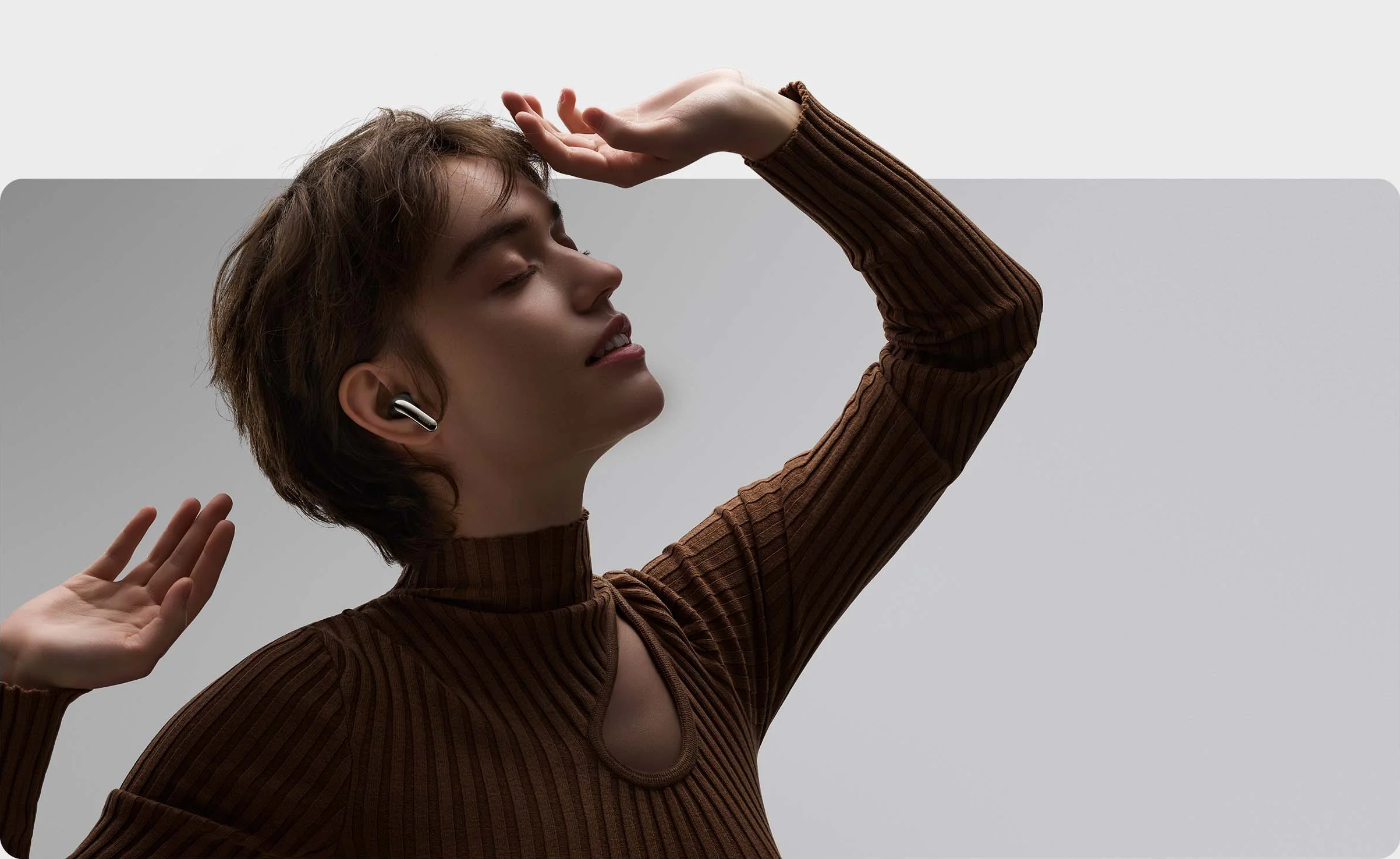 Une femme avec des écouteurs sans fil dans les oreilles, regardant en l'air. Elle porte un pull marron.