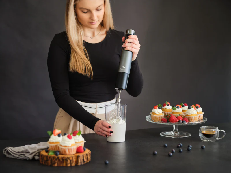 Nő kézi mixerrel tejszínt ver egy pohárban. A háttérben cupcake-ek és bogyók.