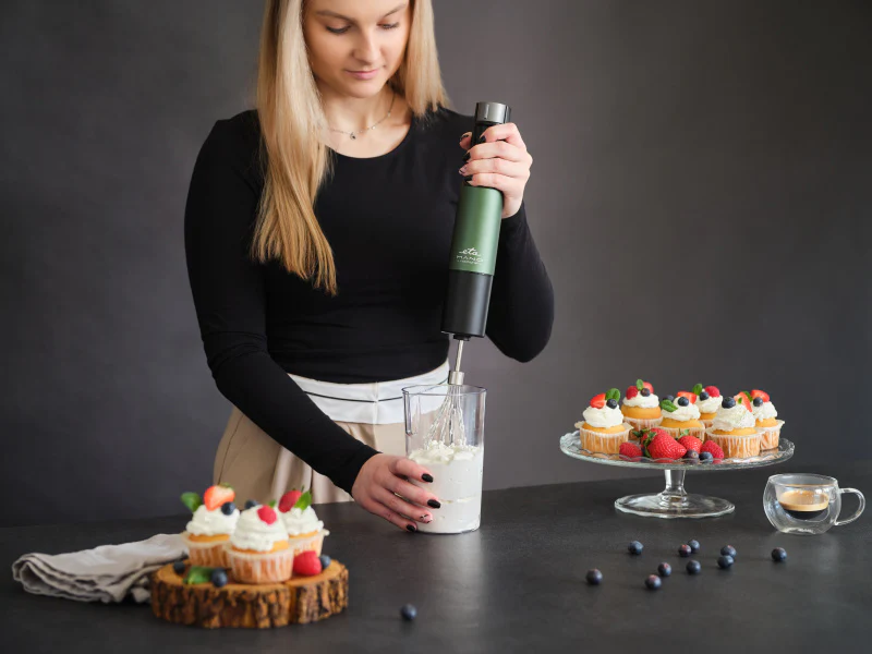 Egy nő kézi mixerrel keveri a krémet egy pohárban, a muffinok mellett.