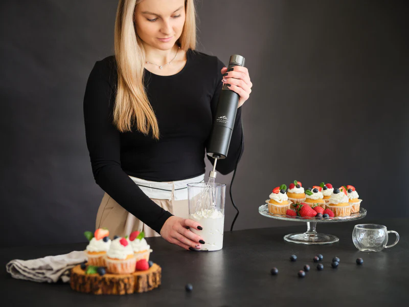 Nő kézi mixerrel tejszínt ver egy pohárban. A háttérben cupcake-ek és bogyók.