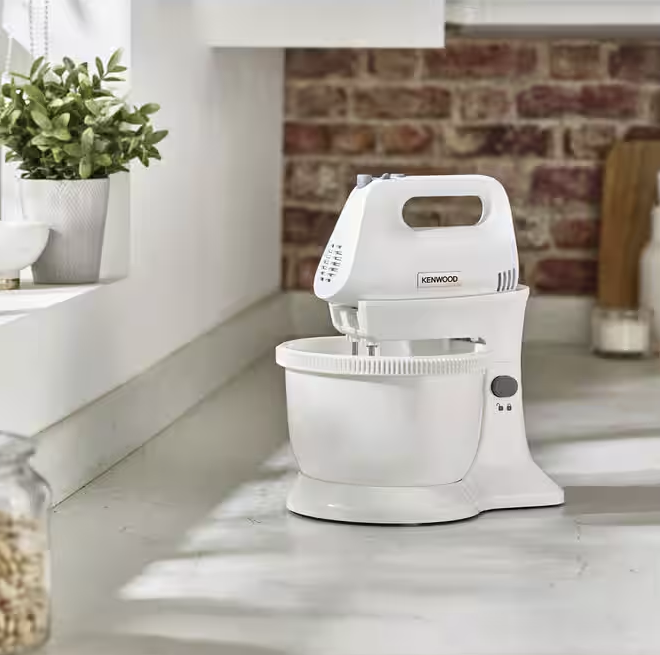 Egy fehér Kenwood robotgép van a pulton. Balra egy növény, a háttérben egy tégl fal.