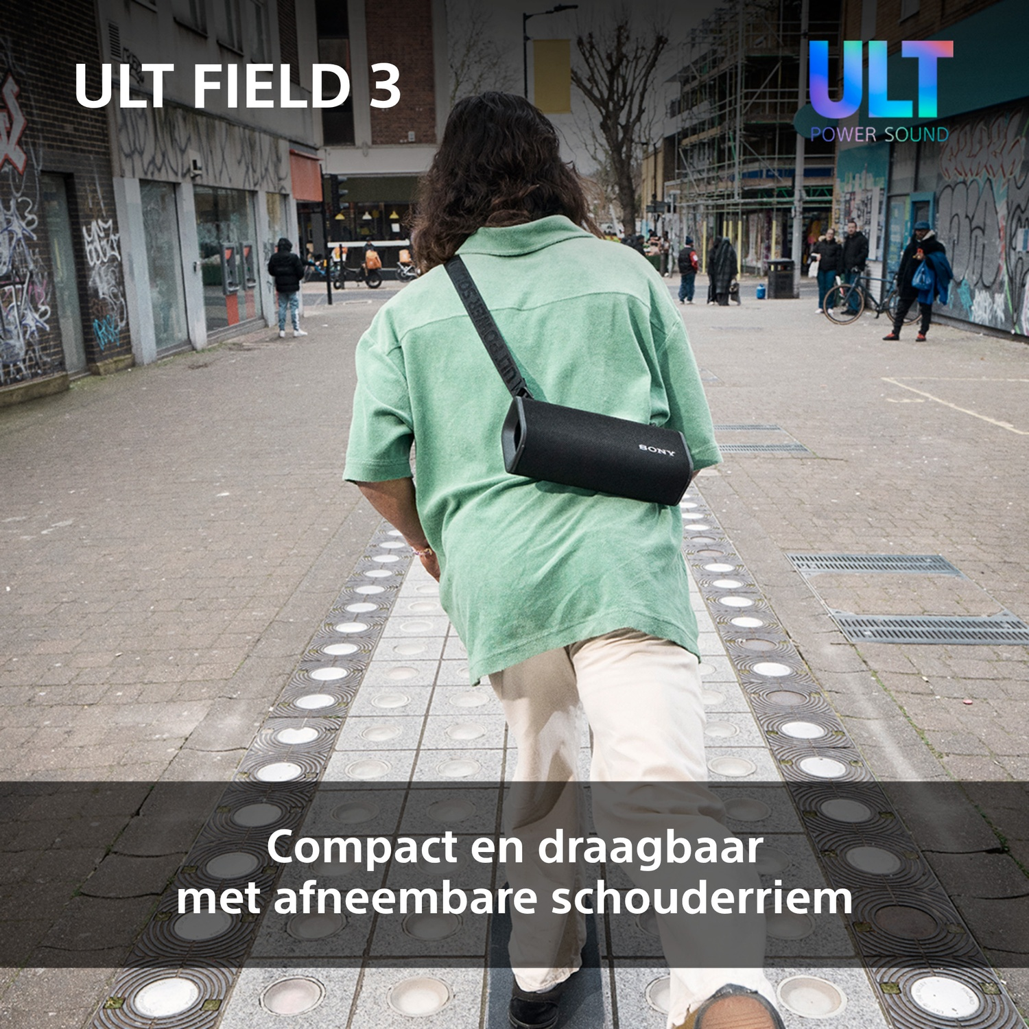 Persoon loopt op straat met een luidspreker. De luidspreker wordt met een schouderriem gedragen. Gebouwen en mensen op de achtergrond.