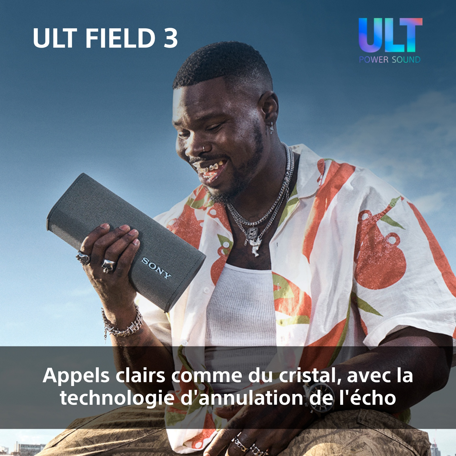 Un homme sourit, tenant un haut-parleur gris. Il porte une chemise colorée, des bijoux. Le ciel est bleu, et le haut-parleur a le logo Sony.