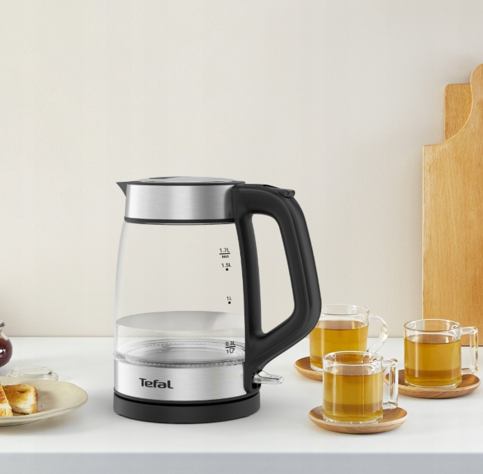 Szklany czajnik z czarnym uchwytem, obok trzech kubków z herbatą i jedzeniem.