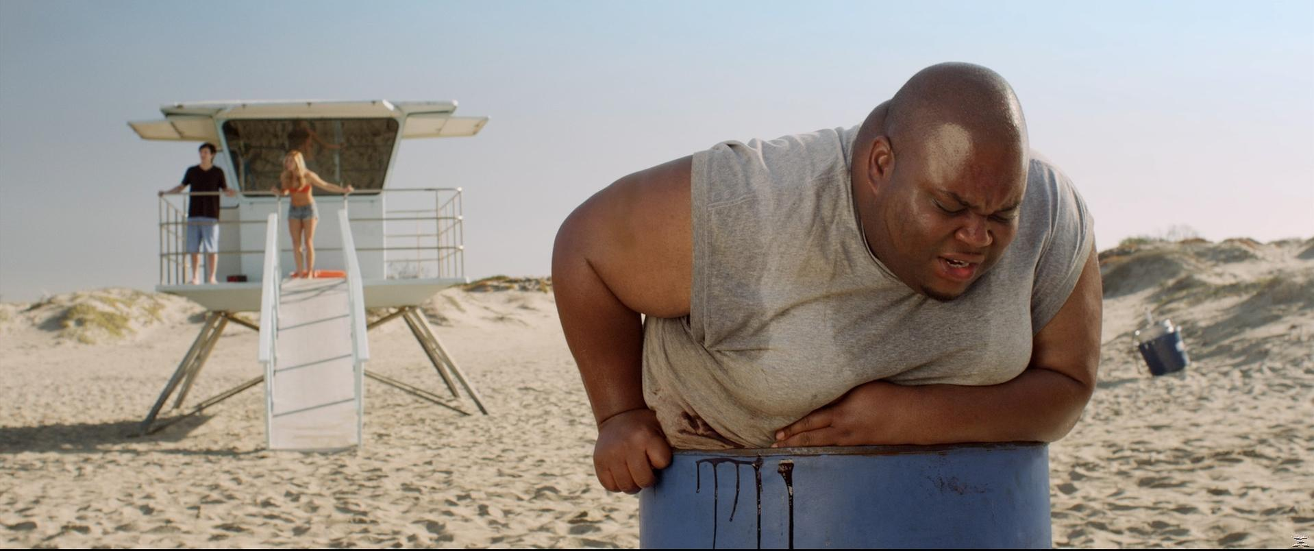 Mann in einem Fass am Strand, im Hintergrund zwei Personen an einem Rettungsschwimmerstand.