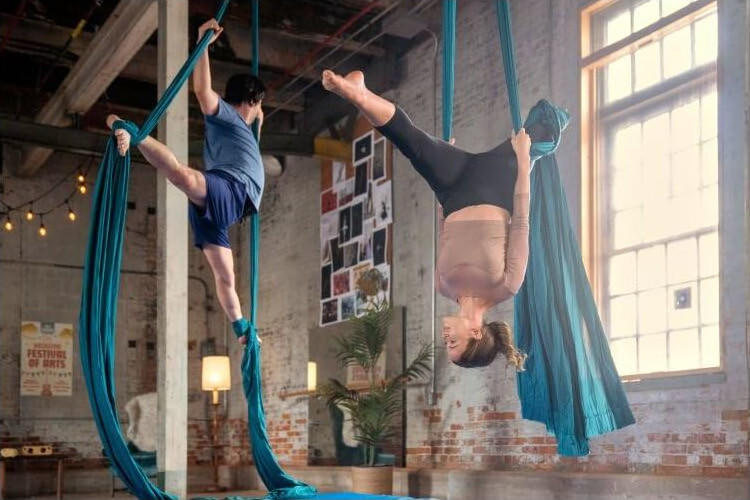 Zwei Personen machen Aerial Yoga. Sie hängen an türkisblauem Stoff in einem Raum.