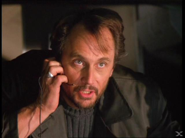 Mann mit Bart telefoniert, blickt nach oben. Dunkler Hintergrund. Er trägt Pullover und Jacke.