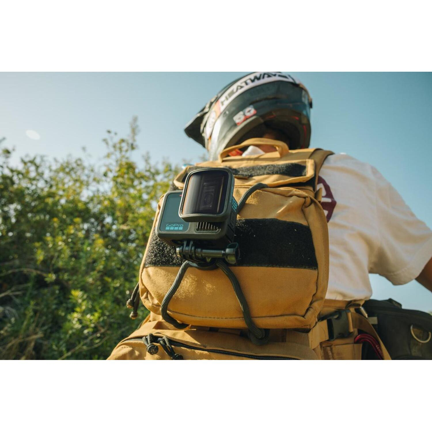 Persoon met helm en GoPro-camera op een rugzak. Groene struiken op de achtergrond, blauwe hemel.