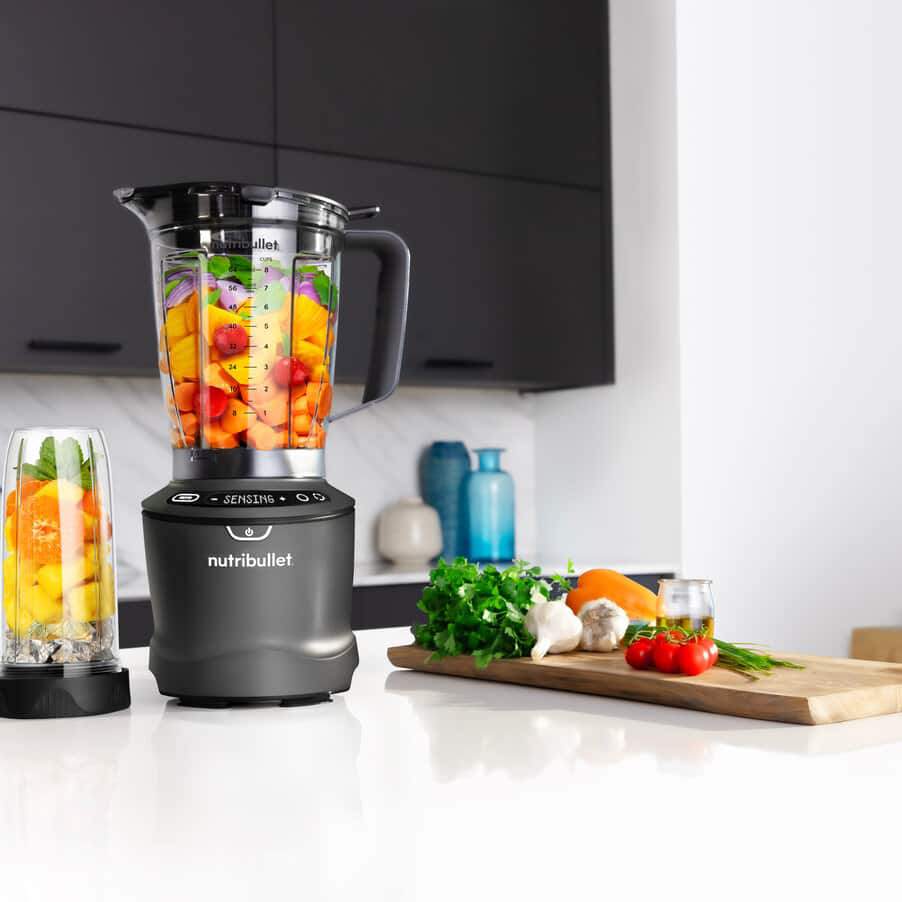 Egy fekete Nutribullet turmixgép tele színes gyümölcsökkel és zöldségekkel, fehér felületen.