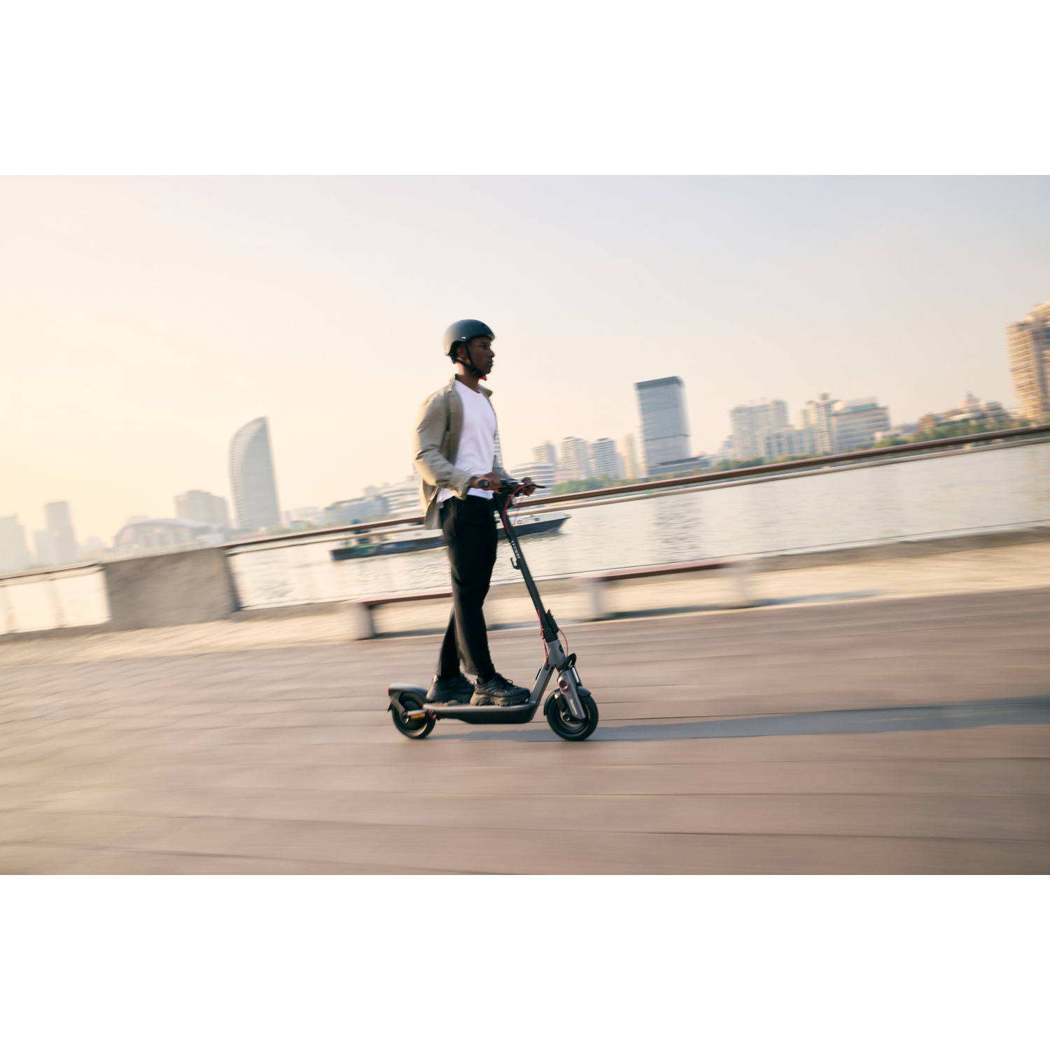 Man riding a grey scooter on a wooden walkway, with water and buildings in the background.