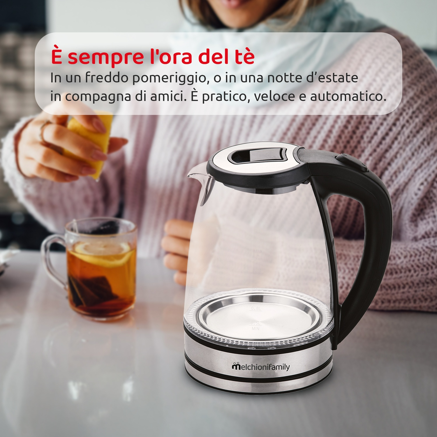 Un bollitore in vetro su base argentata con una persona che tiene un limone, vicino a una tazza di tè.