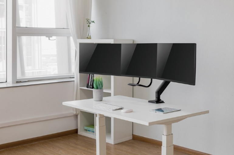 Három számítógép monitor egy fehér asztalon billentyűzettel, egérrel és tablettel egy szobában.