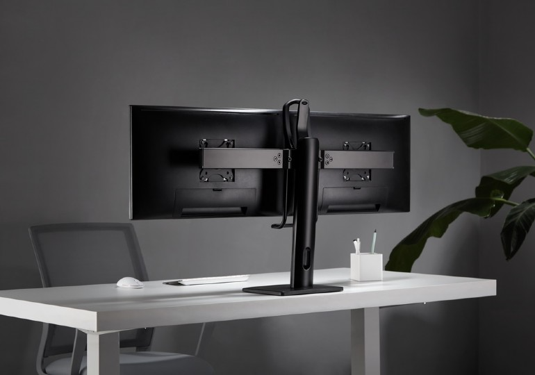 Egy monitor hátulnézete egy fehér asztalon. Háttérben egy növény és egy szürke fal.