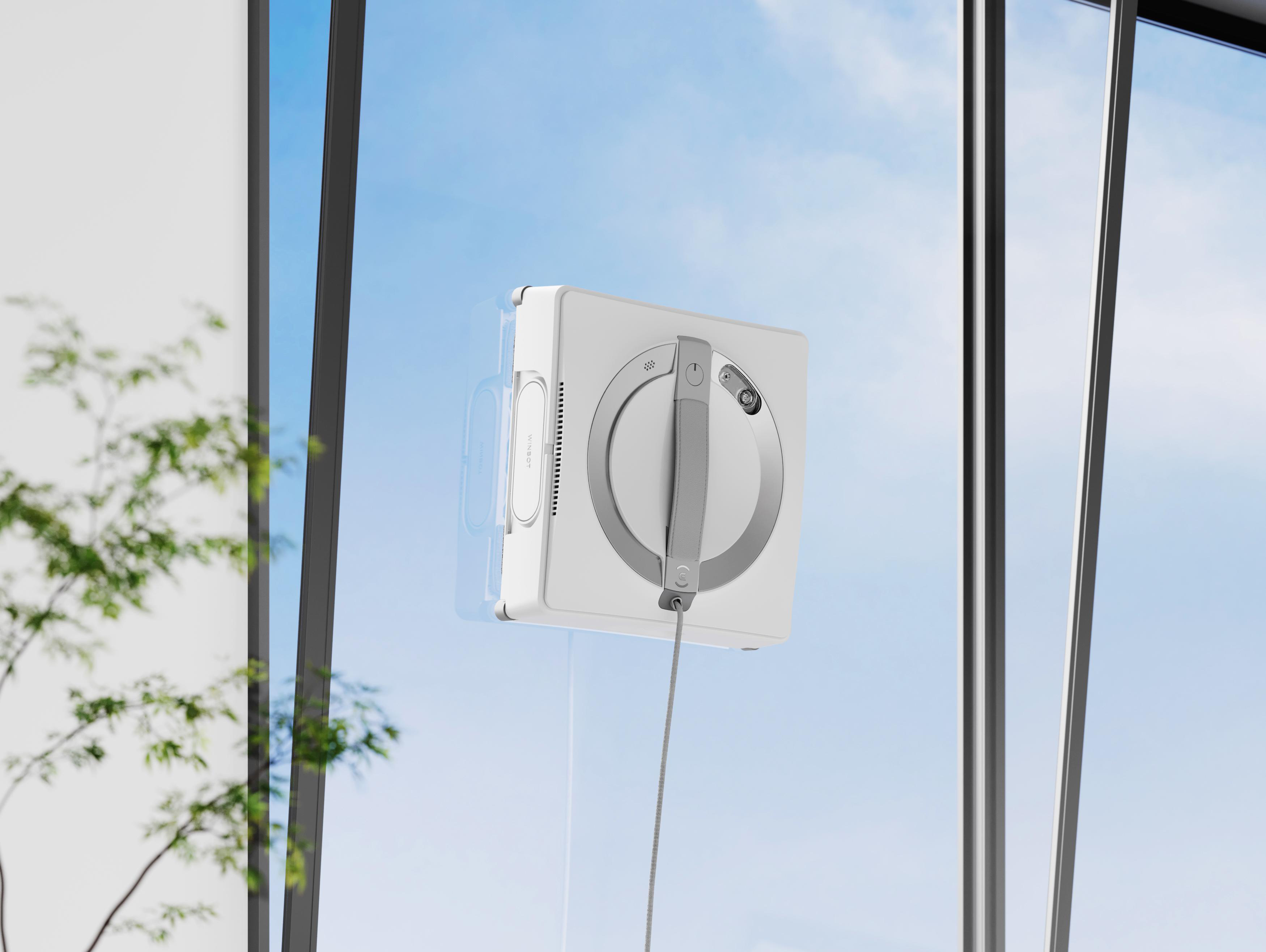 Ein weißer Roboter-Fensterputzer ist an einer Glasscheibe befestigt, durch die ein blauer Himmel zu sehen ist.
