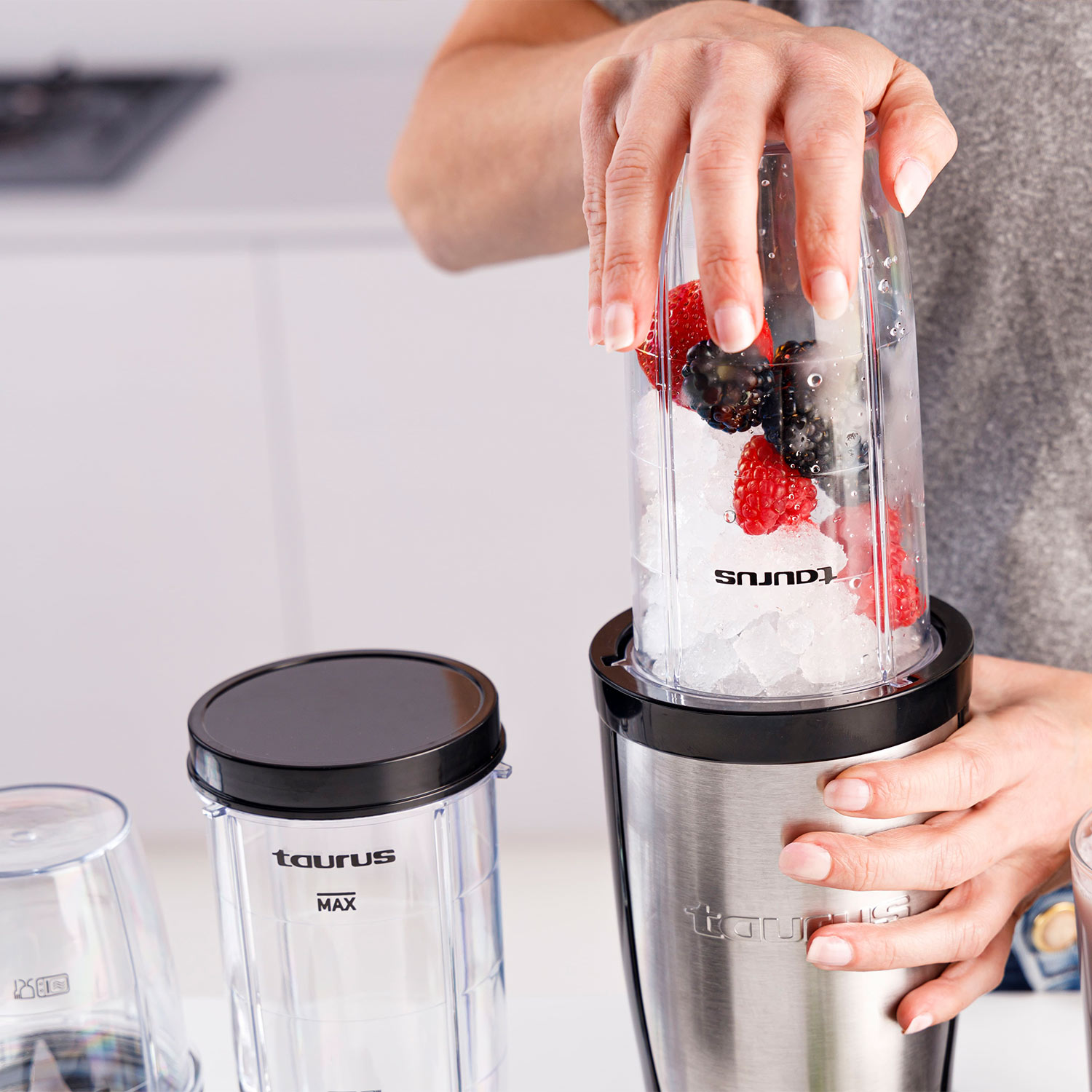 Una persona sostiene una taza de licuadora llena de hielo, bayas y fruta. La taza es de acero inoxidable.