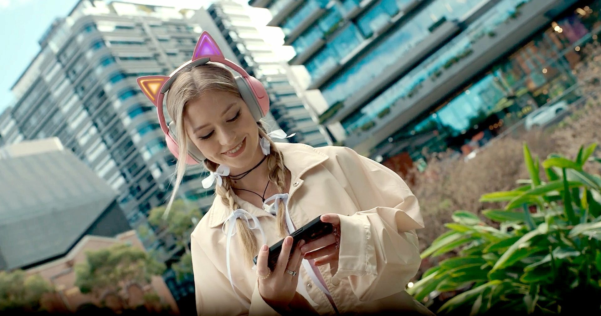 Kobieta ze słuchawkami z uszami kota, uśmiechnięta, trzyma telefon. Miejskie tło.