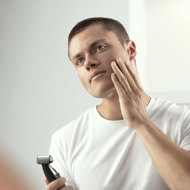 Mężczyzna patrzy w lustro, dotykając policzka dłonią, trzymając golarkę.