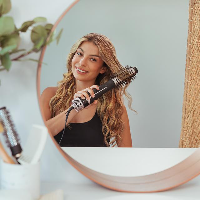 Kobieta stylizuje włosy przed lustrem okrągłą szczotką. Uśmiecha się, na białym tle.
