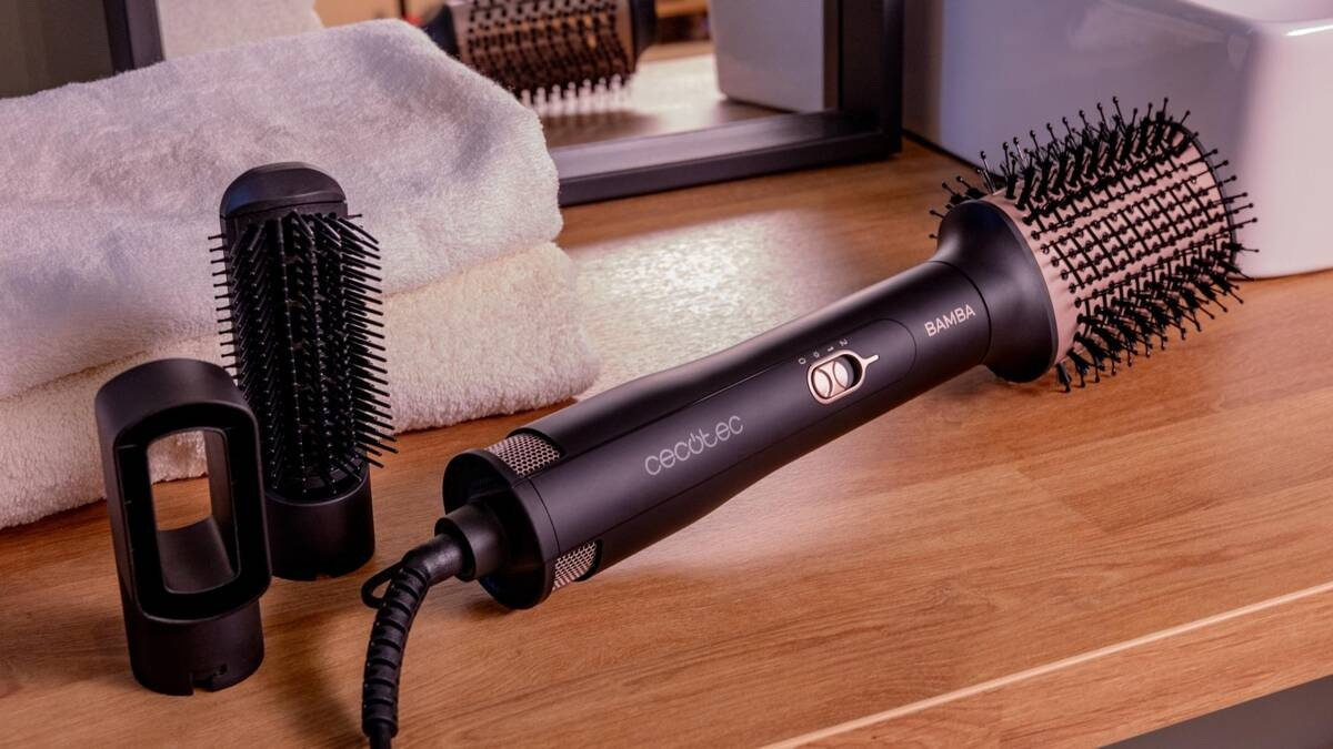 Suszarka do włosów i szczotki leżą na drewnianym stole. Suszarka jest czarna z różowo-złotymi akcentami.
