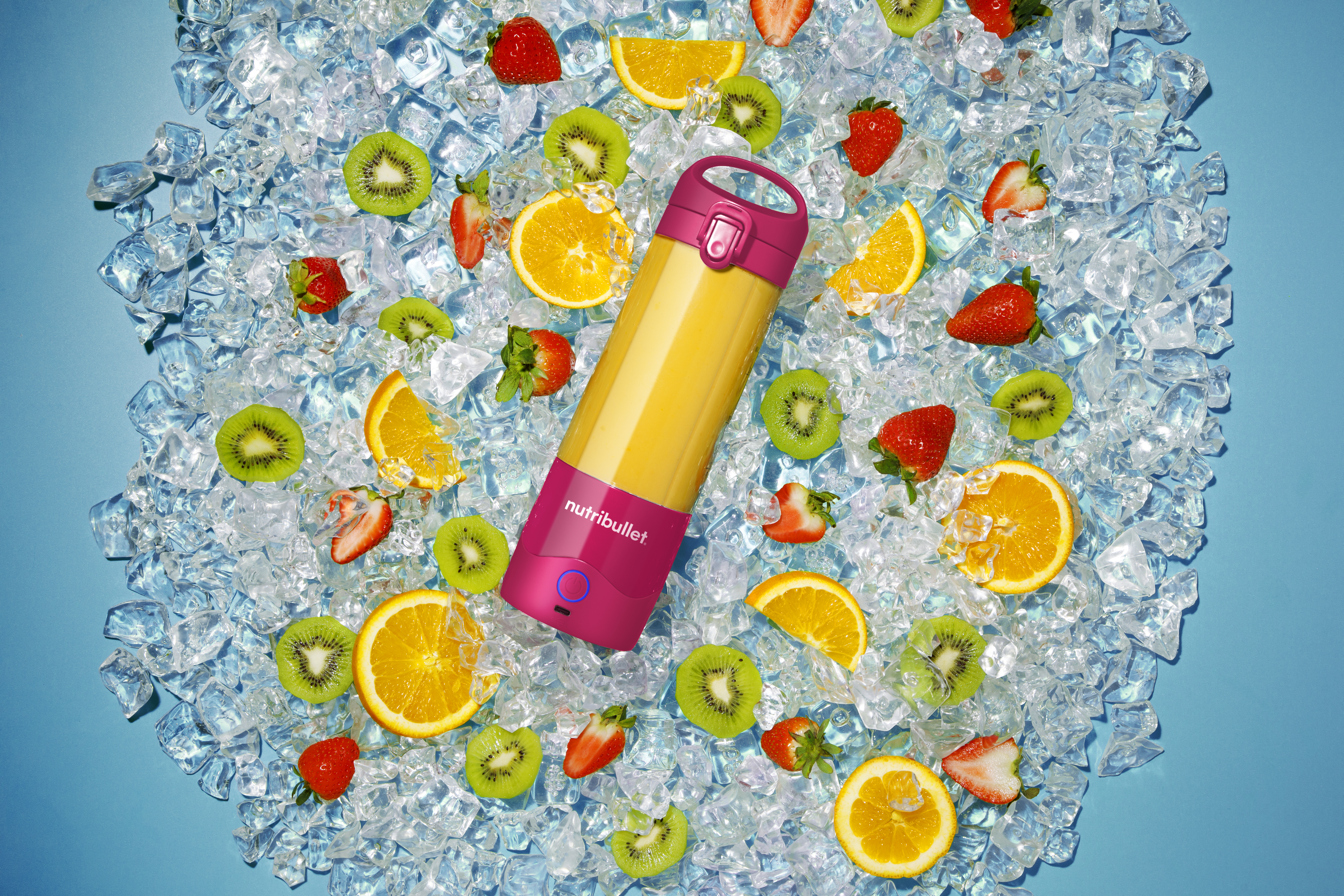 Ein Smoothie-Maker, umgeben von Eis, Kiwi, Orangenscheiben und Erdbeeren auf hellblauem Hintergrund.