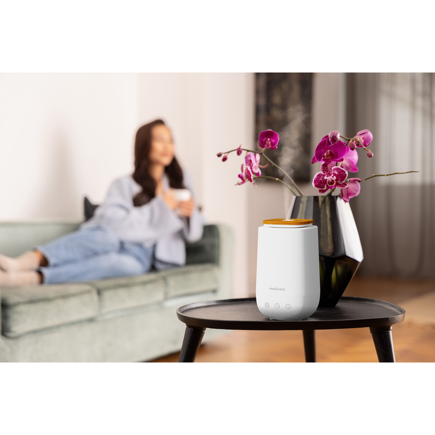 Un diffuseur d'aromathérapie blanc avec un vase d'orchidées roses et une femme sur un canapé.