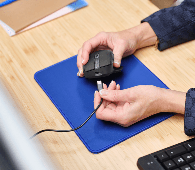 Egy személy egy egeret és egy USB-C kábelt tart egy kék egérpadon egy fa asztalon.