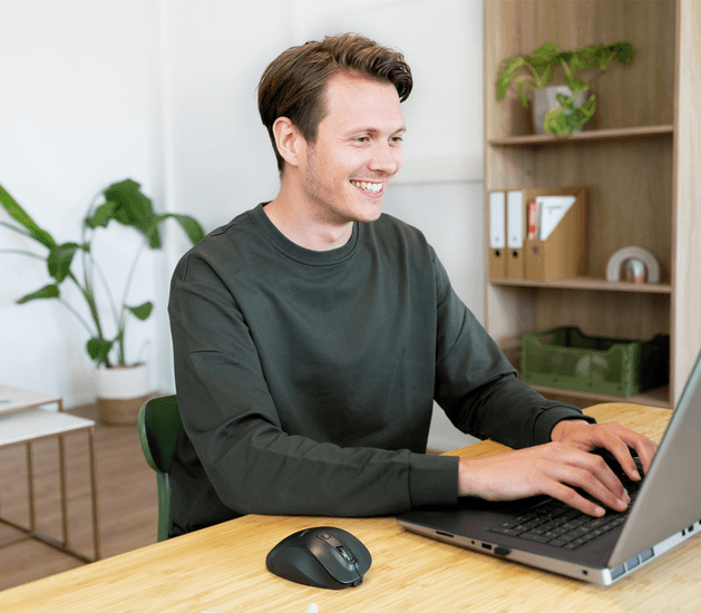 A férfi az asztalnál mosolyog, laptopon gépel. Egér és növény is látható.