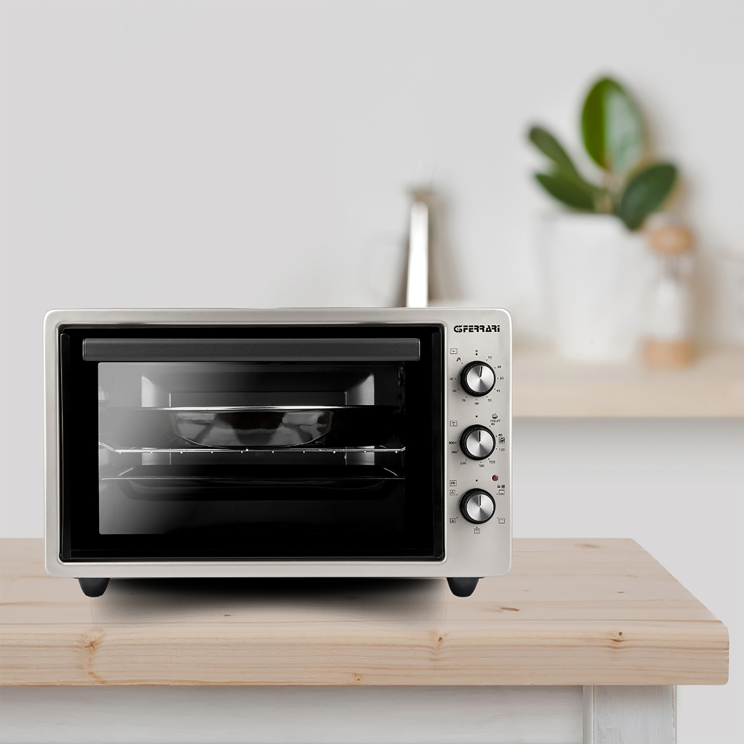 Piccolo forno bianco su superficie in legno con manopole e porta in vetro. Parete bianca e pianta sullo sfondo.