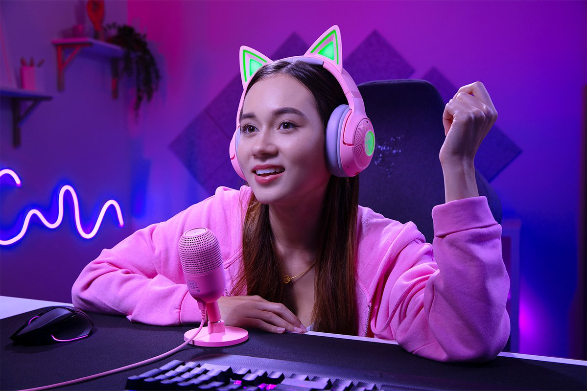 Kobieta ze słuchawkami z kocimi uszami, uśmiechnięta i grająca z różowym mikrofonem i klawiaturą.