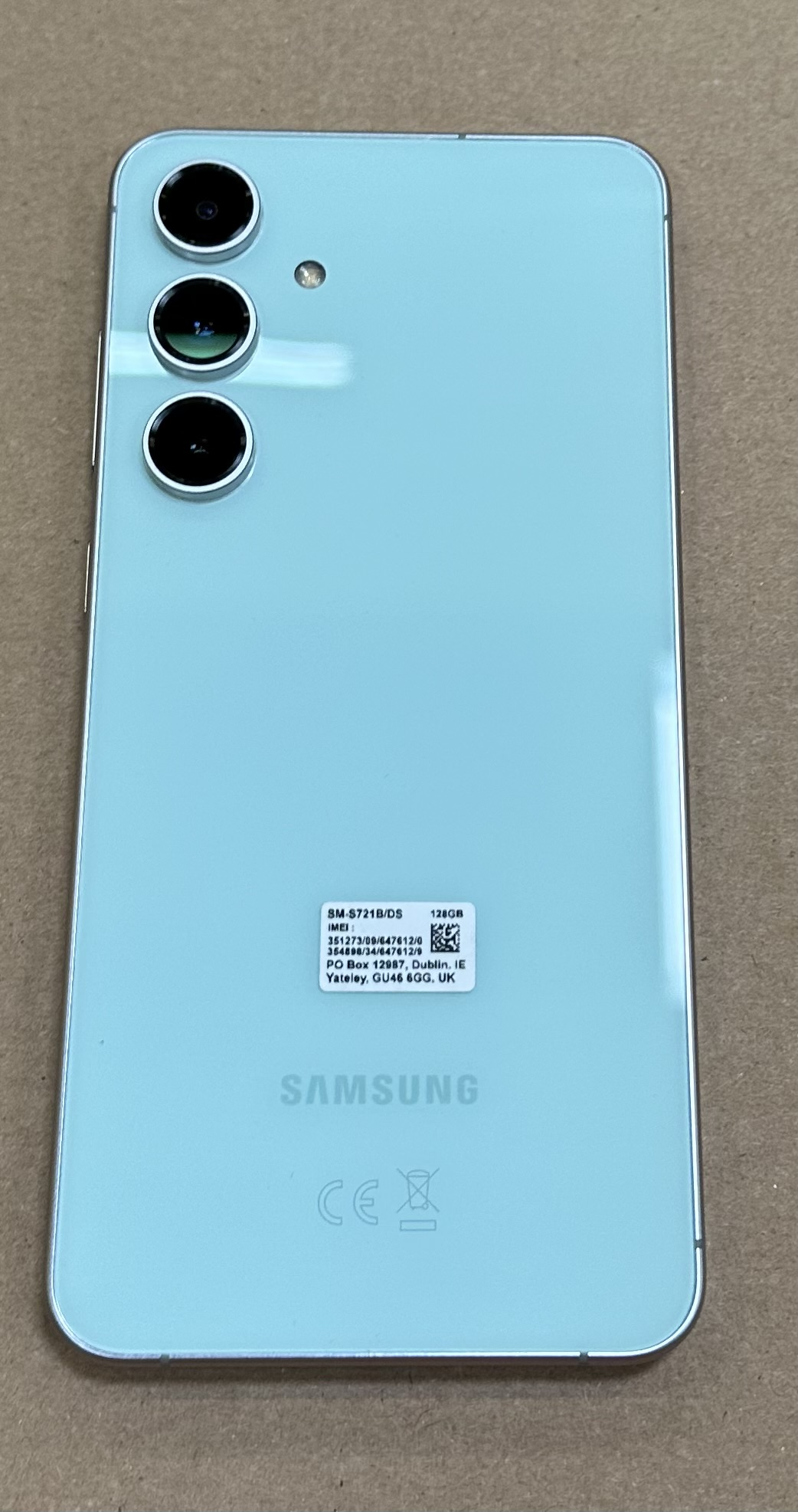 Jasnoniebieski telefon Samsung z trzema tylnymi aparatami, na brązowej powierzchni. Telefon ma naklejkę.