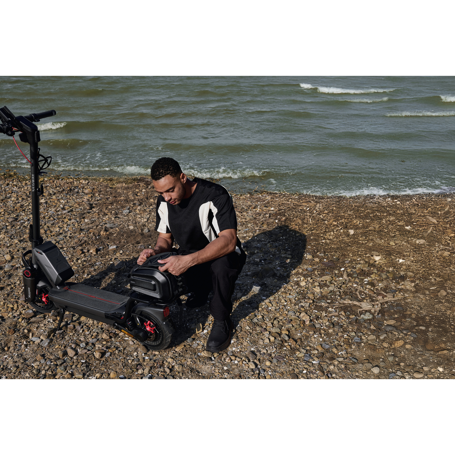 Man knielend bij een zwarte scooter op een rotsachtig strand. Oceaan op de achtergrond, groen water.
