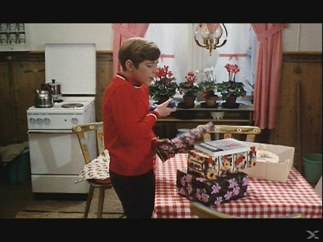 Ein Junge in einem roten Pullover mit Geschenken in einer Küche mit karierter Tischdecke.