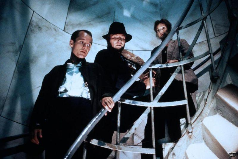 Drei Personen auf einer Wendeltreppe. Einer hält eine Geige. Die Szene ist schwach beleuchtet.