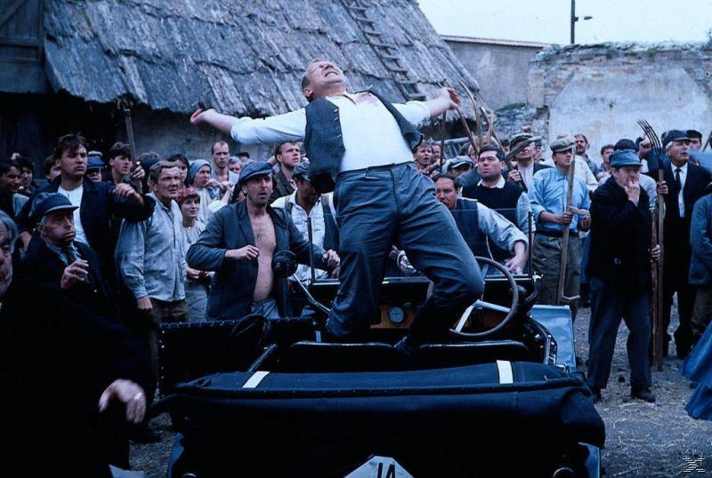 Mann auf Auto, Arme hebend, umgeben von Leuten. Szene aus einem Film in einem Dorf.