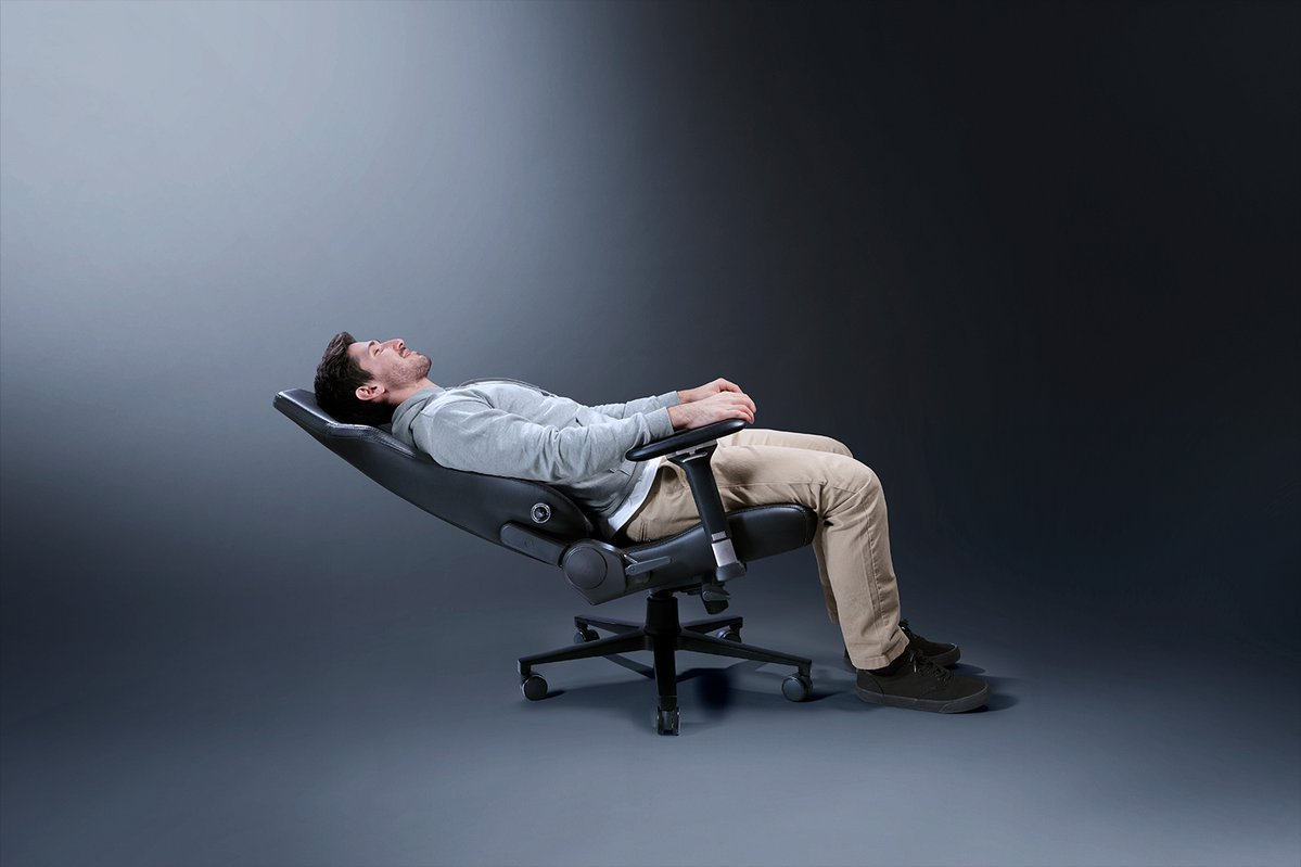 Mężczyzna leżący w czarnym ergonomicznym krześle, głowa do tyłu, w studio z szarym tłem.