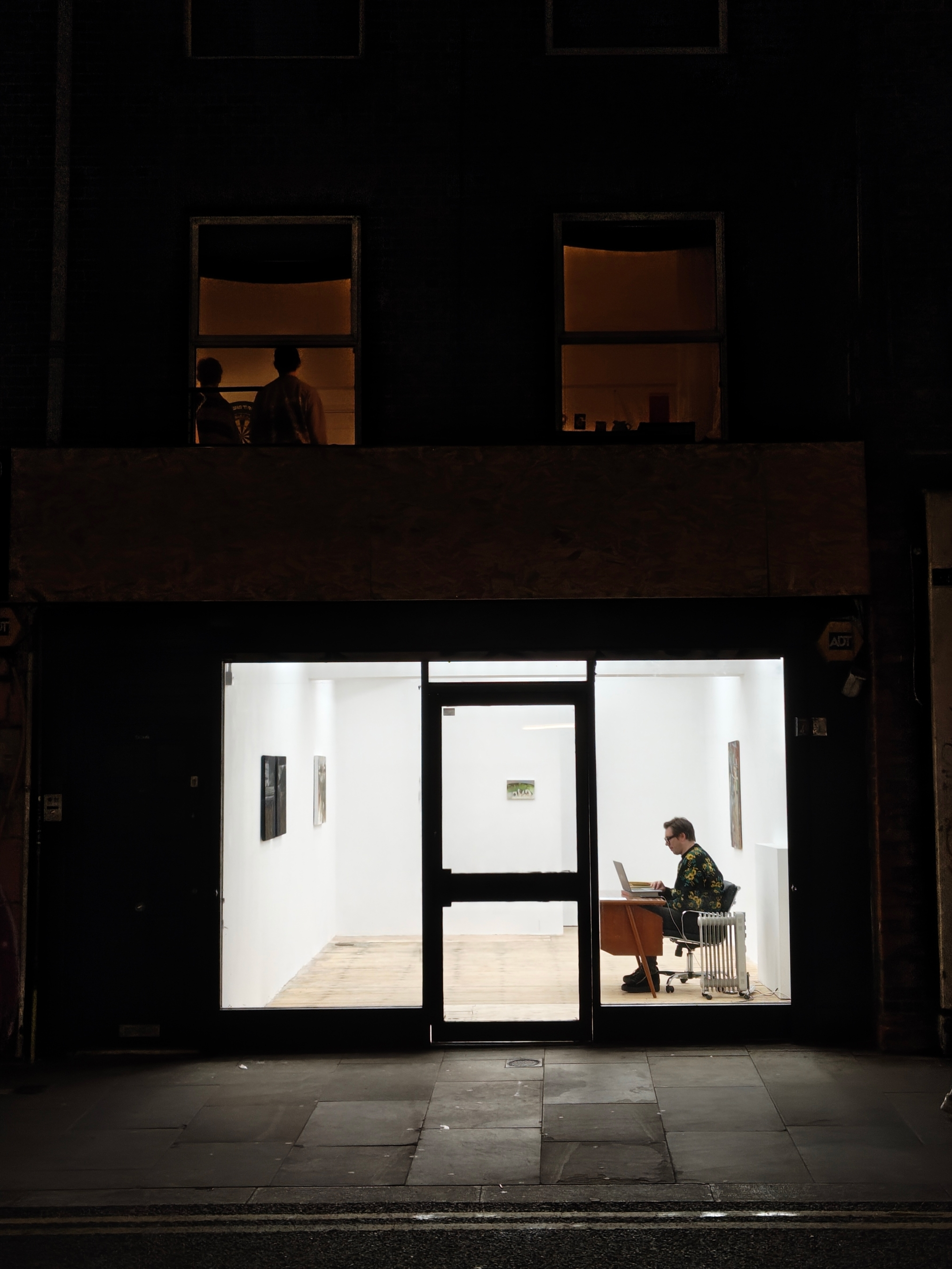 Vue extérieure d'une galerie la nuit. Des personnes sont visibles dans les fenêtres supérieures ; un homme travaille à l'intérieur.