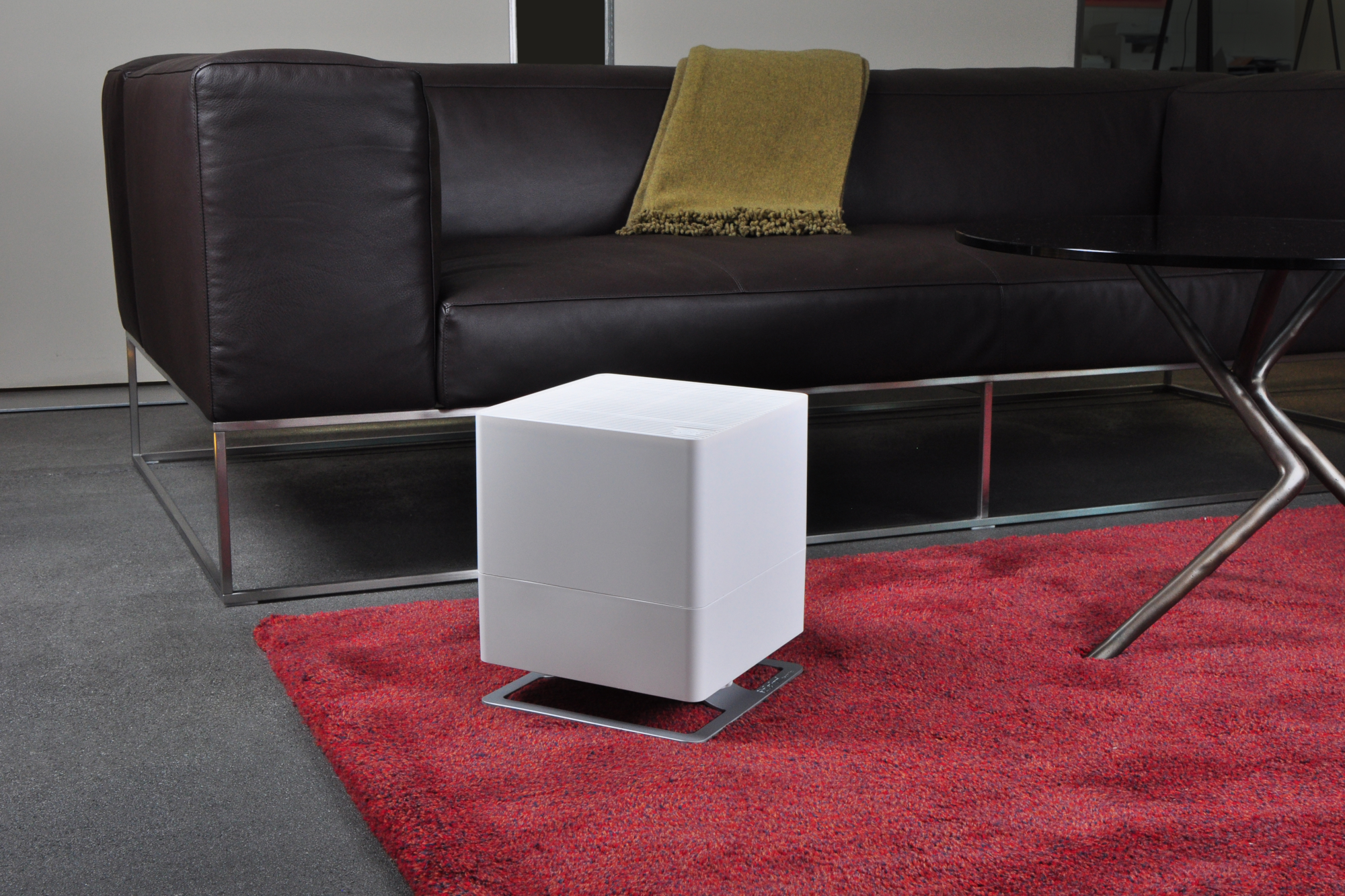 Purificatore d'aria bianco su un tappeto rosso, con divano in pelle marrone e tavolo nero sullo sfondo.